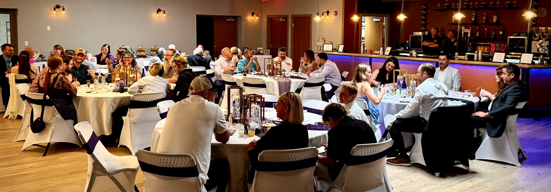 Wedding reception with guests seated at round tables beside a blue-lit bar